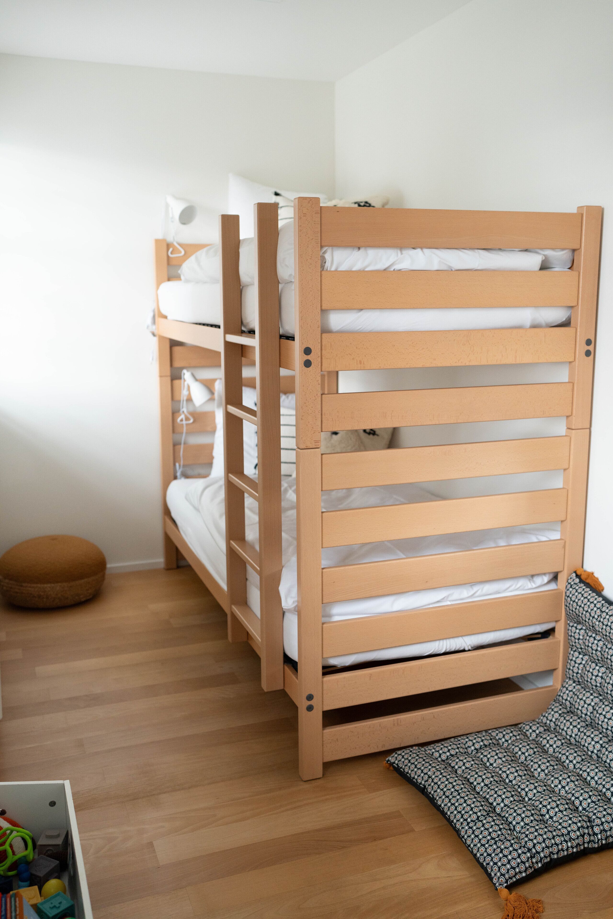 Ein Etagenbett mit Holzrahmen und weißer Bettwäsche, ein runder Hocker und ein gemustertes Teppich auf Holzdielen.