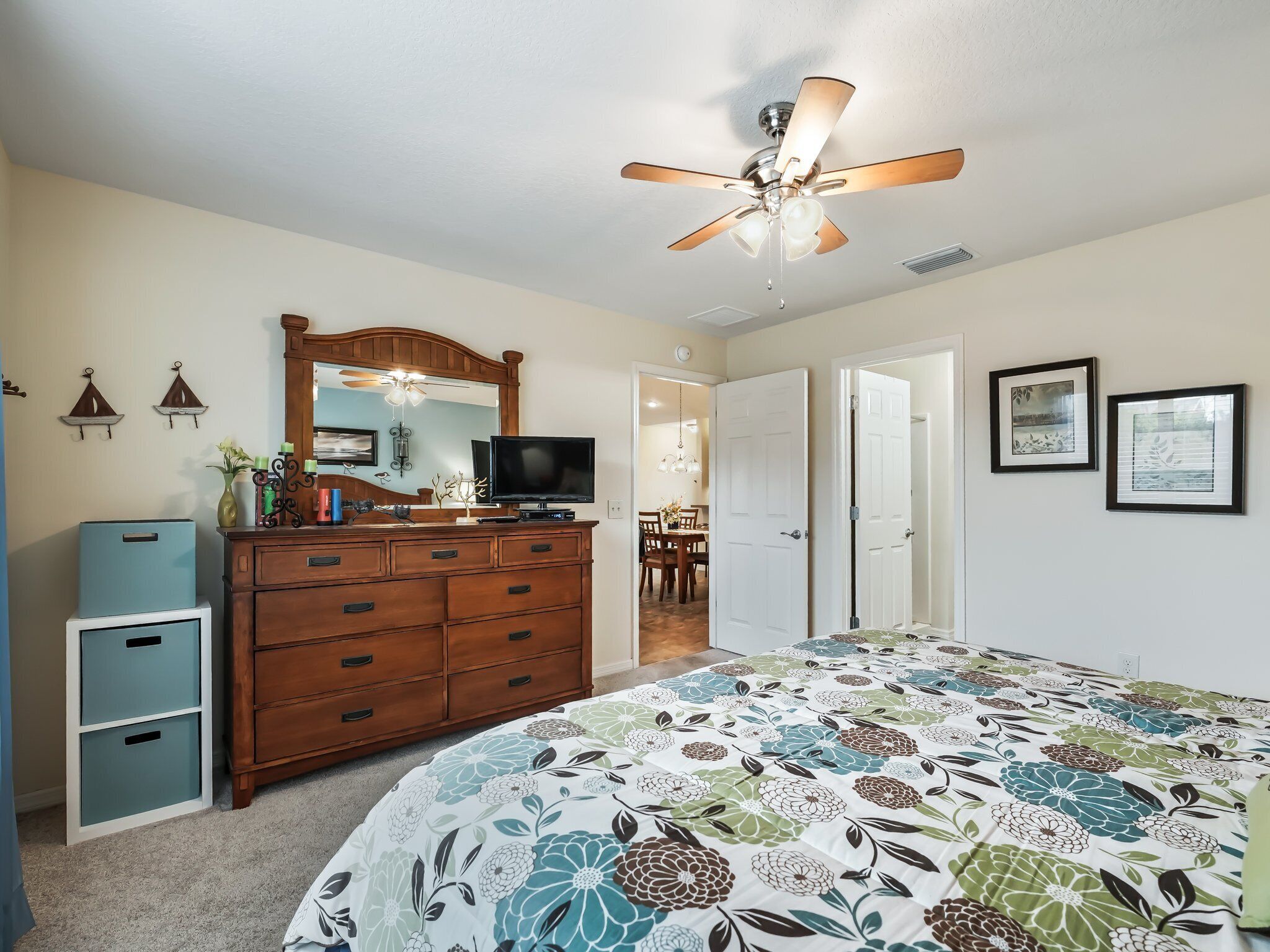 Une chambre à coucher comprenant un lit, une commode, un téléviseur, un ventilateur de plafond et une porte donnant sur une autre pièce.