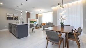 A modern kitchen with a central island, a dining area with a wooden table and chairs, and a living space with a sofa and a TV. - 4BR/3BA Disney-Themed Townhome at Storey Lake (Kissimmee)