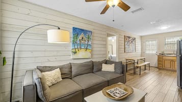 Un salon comprenant un canapé d’angle, une table à manger, un ventilateur de plafond et un coin cuisine avec des meubles en bois.