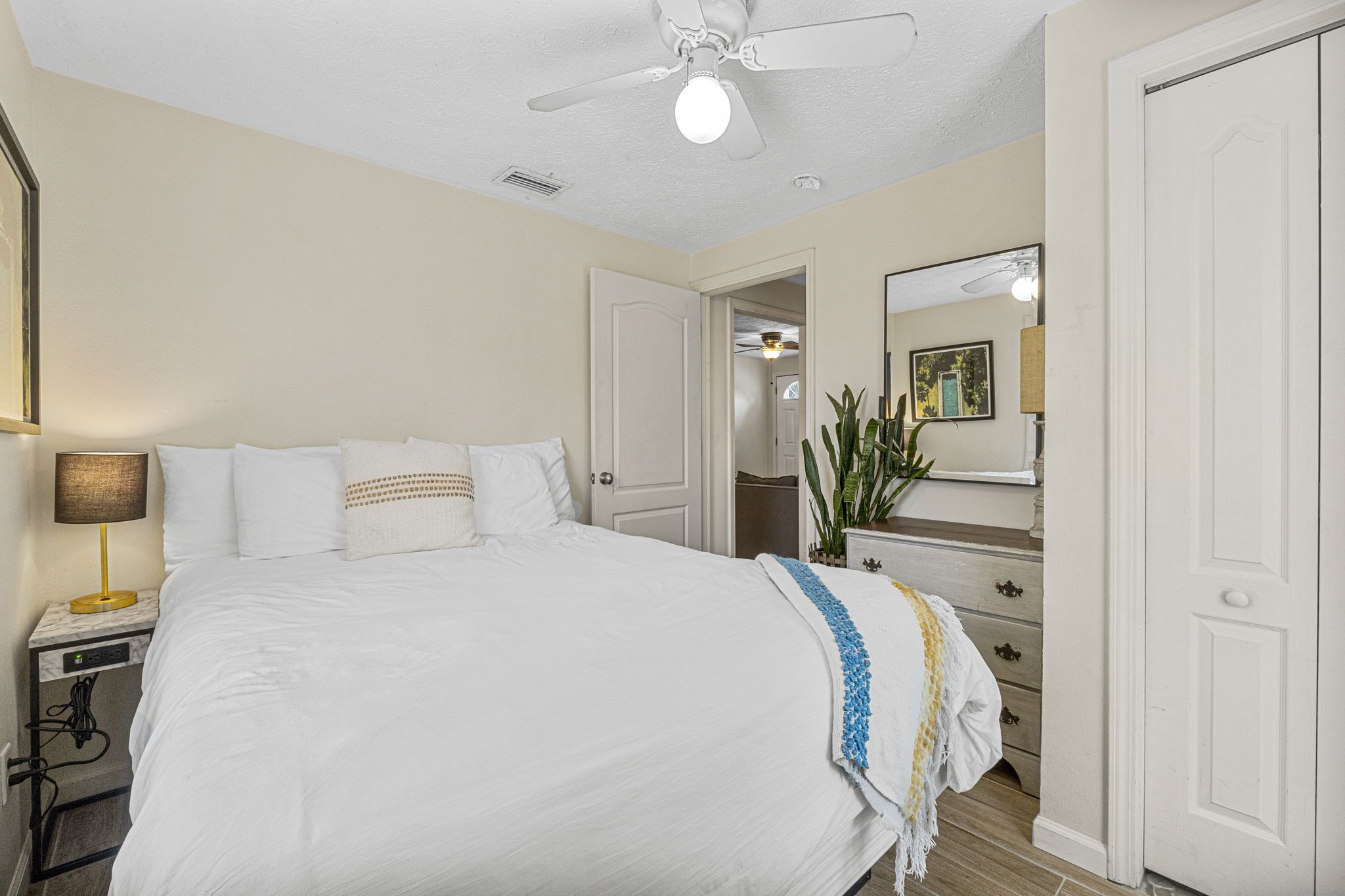 Une chambre d’hôtel comprenant un lit, une table de chevet, une lampe, une commode, un miroir et un ventilateur de plafond.