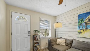 Un salon avec une porte beige, une bibliothèque, un canapé gris et un tableau représentant une scène de plage.
