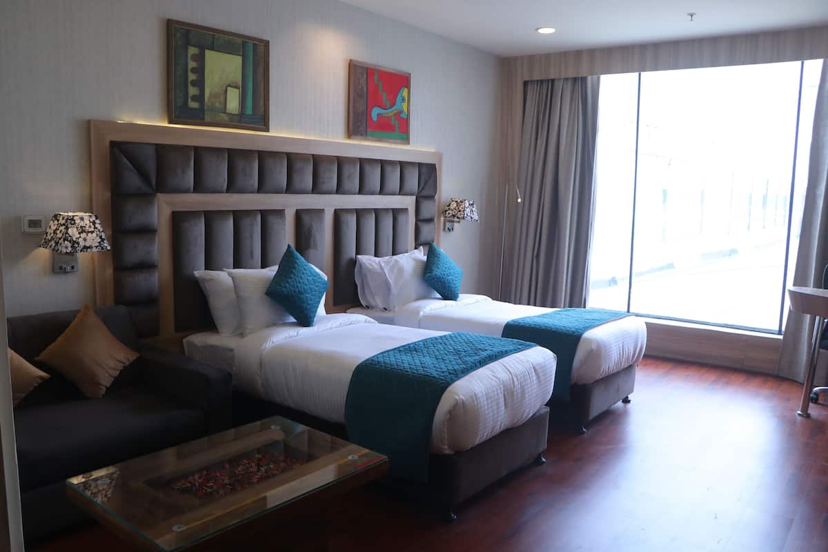 Quarto de hotel com duas camas, um sofá, uma mesa de centro de vidro e uma janela grande com cortinas.