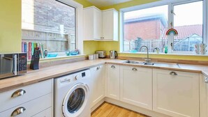 A modern kitchen with white cabinets, a washing machine, a microwave, and a large window with a view of a house and a rainbow decoration. - Host & Stay | St Mark's Close (Marske-by-the-Sea)