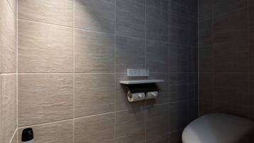 Ein modernes Badezimmer mit grauen Fliesen, einer Toilette, einem Ablagefach mit Pflegeprodukten und einer Leuchte.