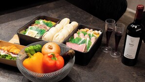 Een eettafel met maaltijdverpakkingen, een schaal groenten en twee wijnglazen met een fles wijn.