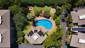 An aerial view of a residential area with a swimming pool surrounded by lush greenery, solar panels, and a small deck area.