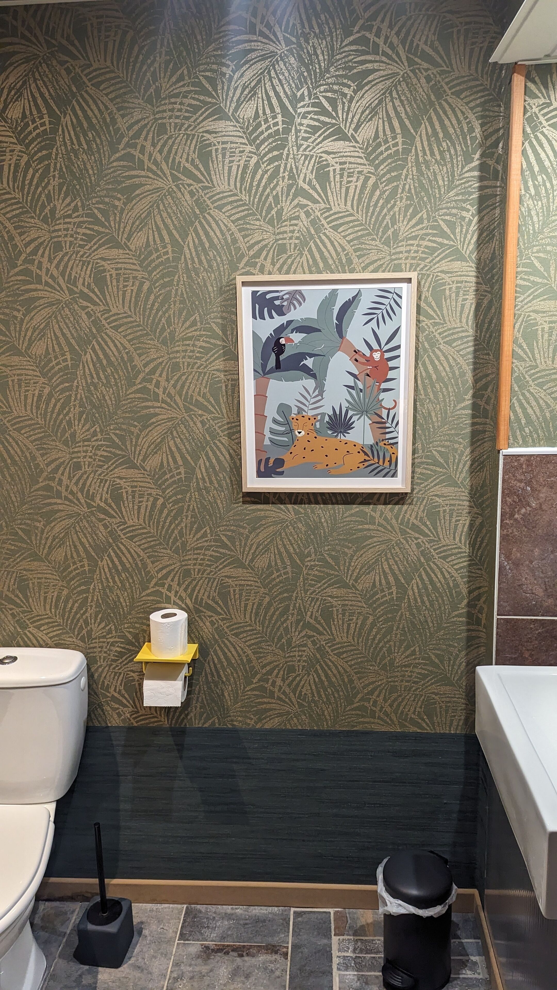 Ein Badezimmer mit einer Wand mit grünem Blattmuster, einer gerahmten Tierillustration, einer Toilette, einer kleinen Ablage mit einer Toilettenpapierrolle und einem Mülleimer.
