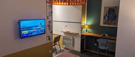 A modern hotel room with a mounted TV displaying a streaming service, a desk with a lamp, a chair, and a decorative wall piece.