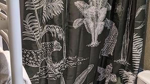 Une salle de bain avec un rideau de douche vert orné de motifs tropicaux blancs représentant des feuilles et des fleurs, des toilettes blanches et un porte-serviettes blanc.