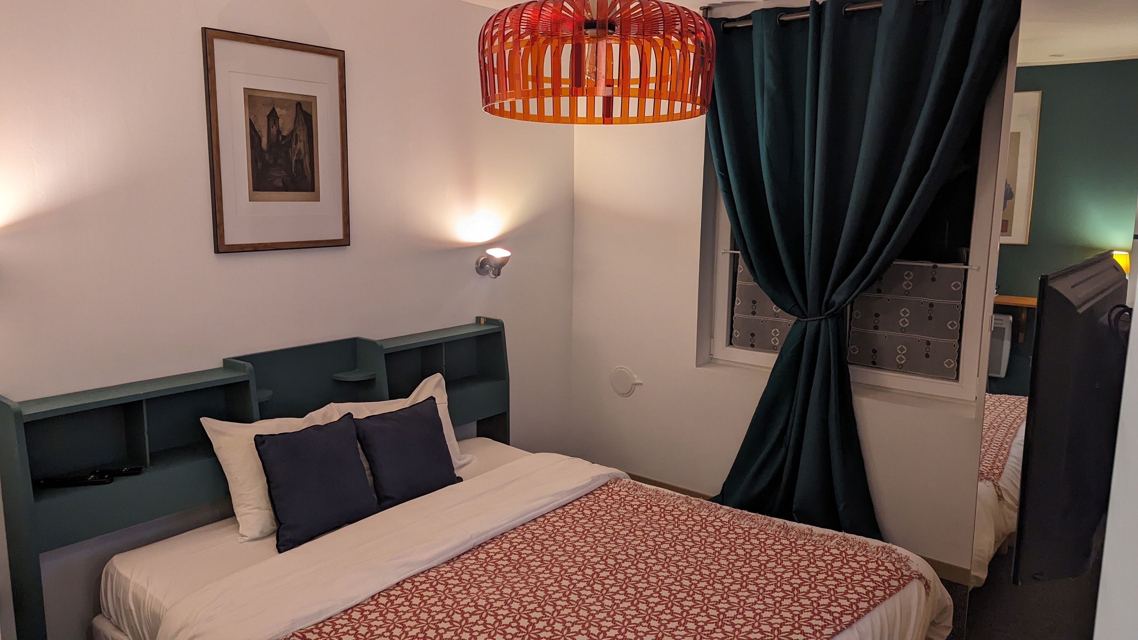 A neatly made bed with a patterned bedspread, a framed picture on the wall, a red hanging light fixture, and a window with curtains.