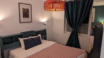 A neatly made bed with a patterned bedspread, a framed picture on the wall, a red hanging light fixture, and a window with curtains.
