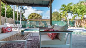 A covered outdoor seating area with a pool, white furniture, and a view of a green fence and trees. - Pompano Oasis w/ Heated Pool 8 mins to the Beach! (Pompano Beach)