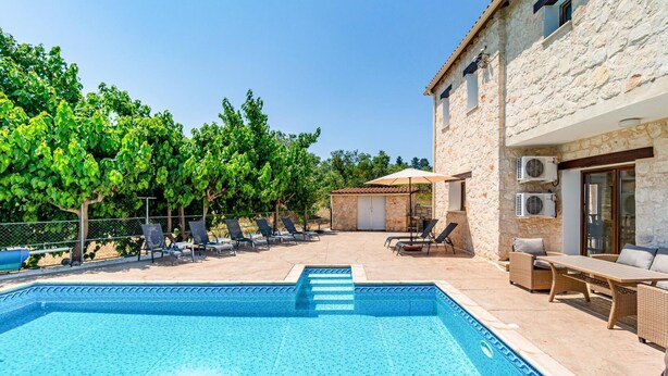 Una piscina con solárium empedrado, sillones de descanso y un edificio de piedra con equipos de aire acondicionado.