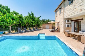 Une piscine dotée d’une terrasse carrelée, de chaises longues et d’un bâtiment en pierre équipé de climatiseurs.