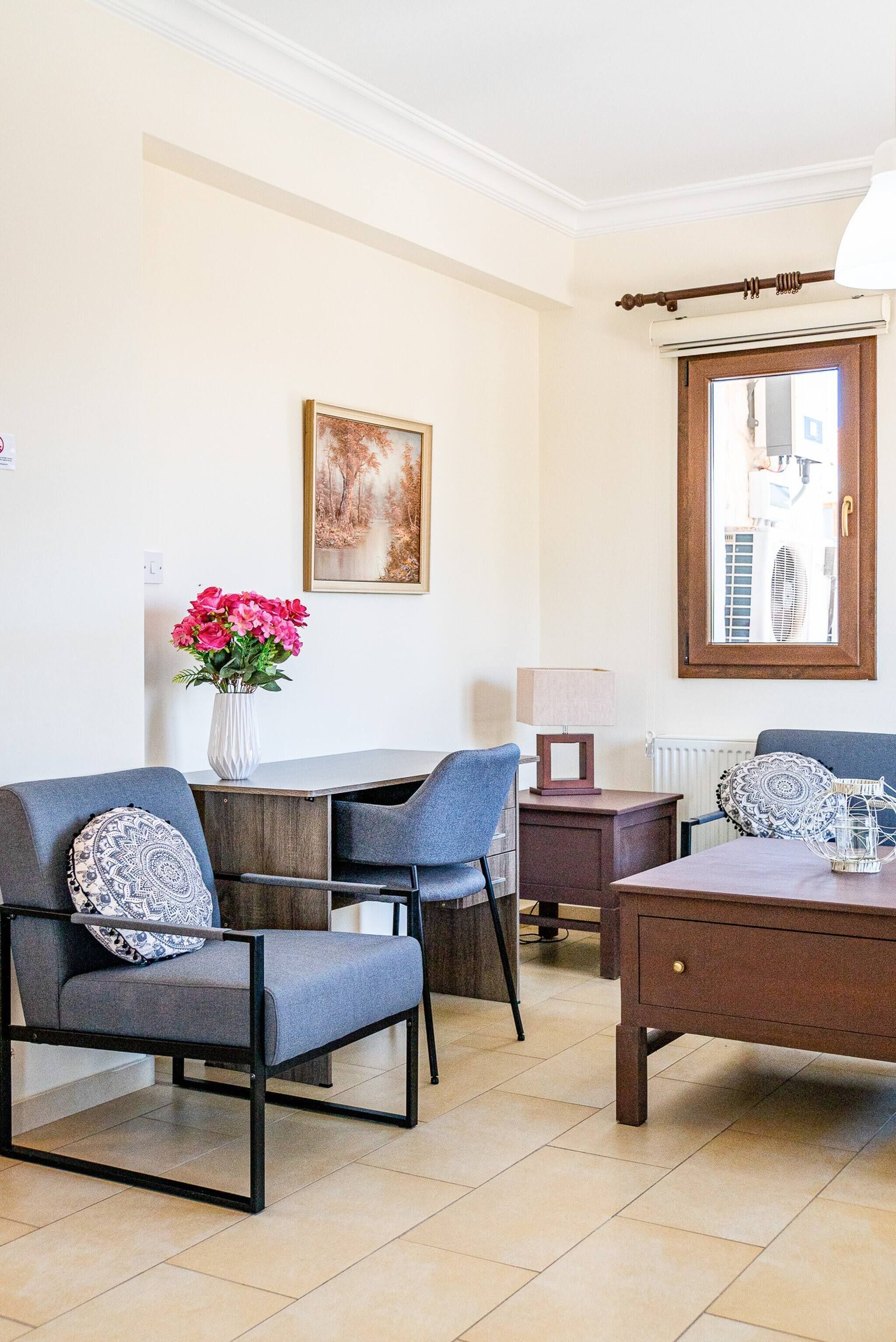 Ein modernes Wohnzimmer mit Esstisch, zwei grauen Stühlen, einem Couchtisch, einem Blumenstrauß, einer Lampe und einem gerahmten Bild an der Wand.