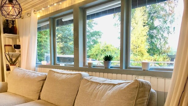 Een beige bank met een gebreide plaid, voor een raam met gordijnen, en een plank met decoratieve objecten.