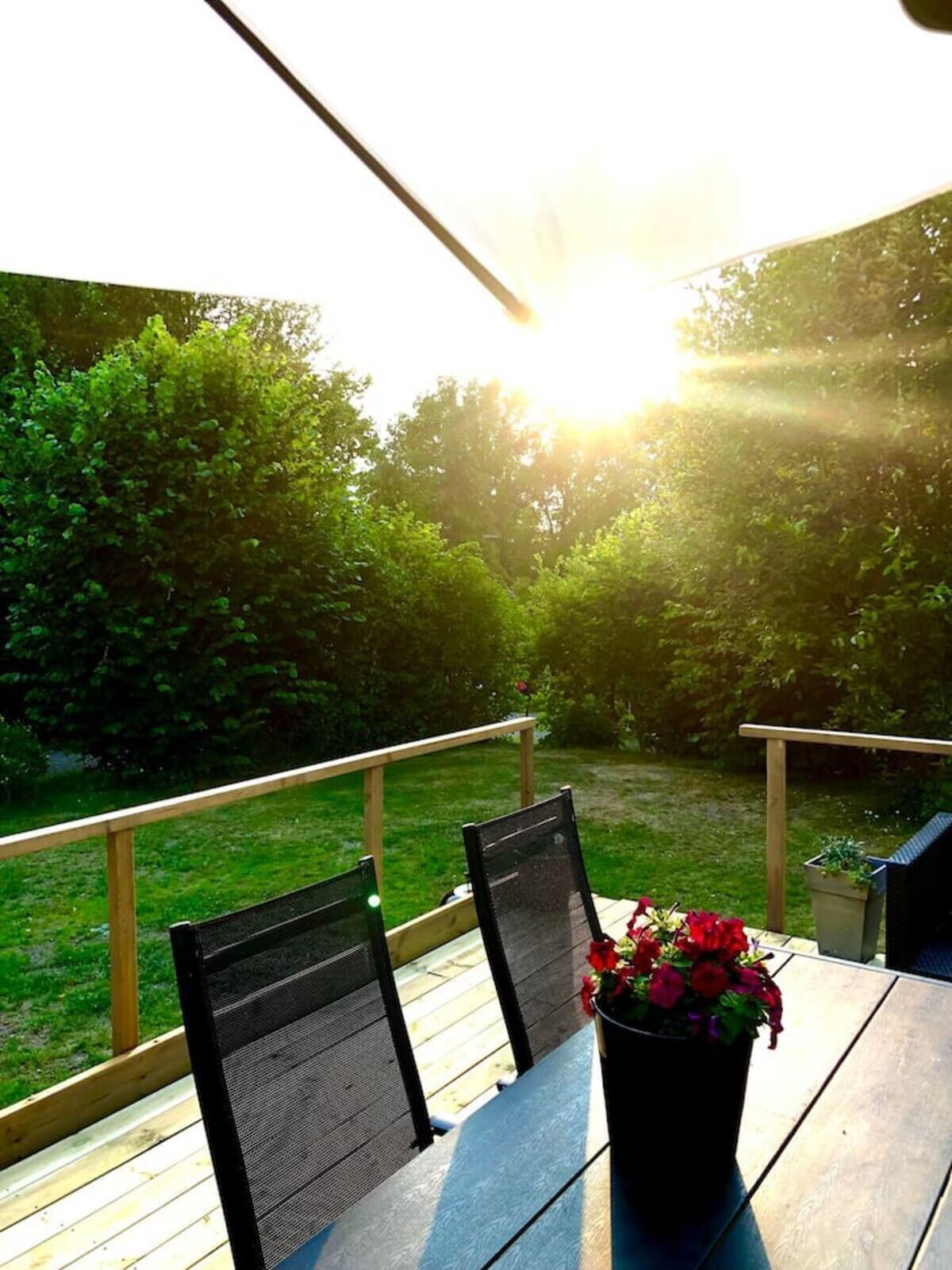 A wooden deck with two chairs and a table, a potted plant with red flowers, and a view of a green garden with trees and a wooden fence.