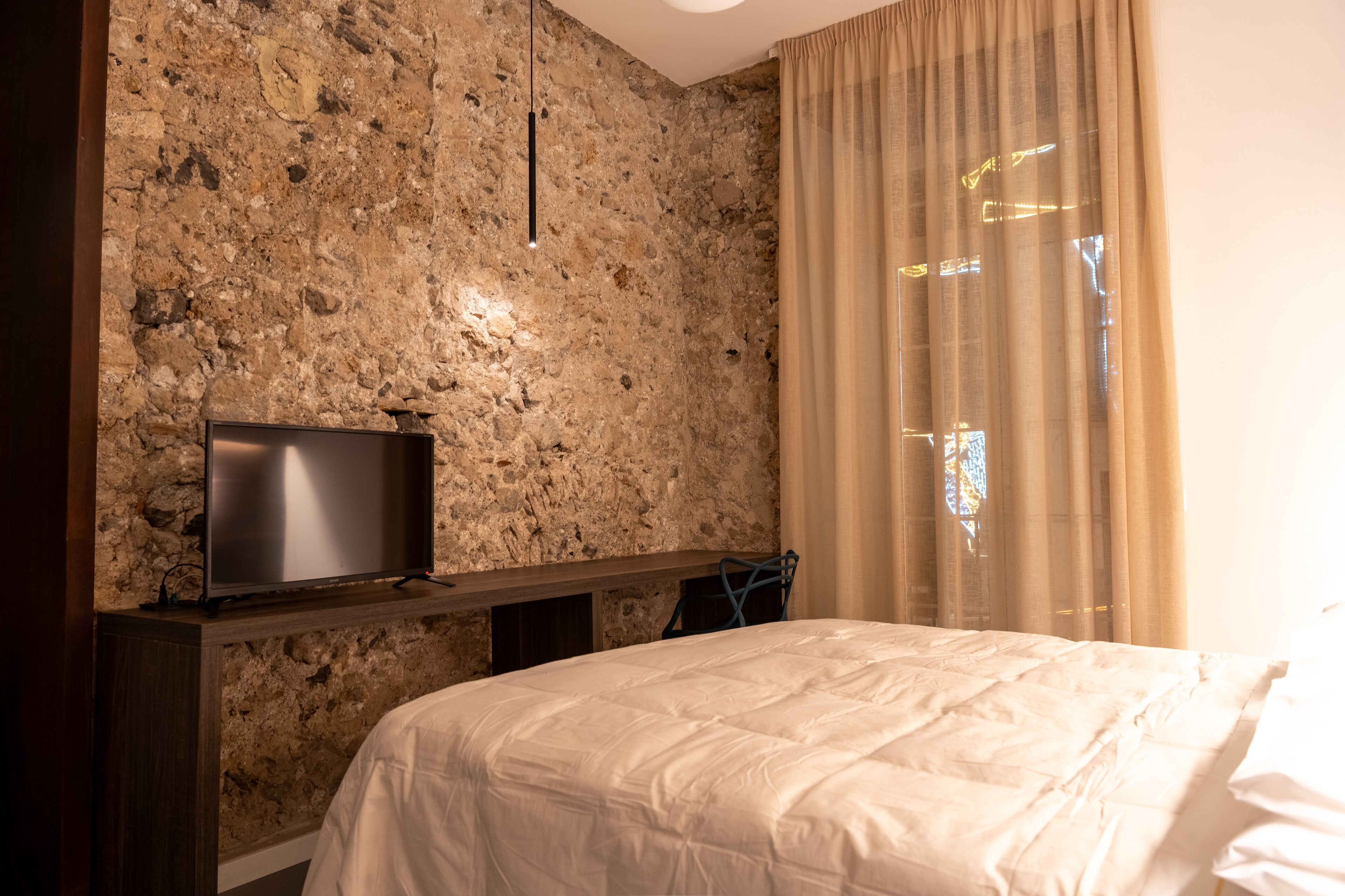 A bedroom with a stone wall, a flat-screen TV, a bed with white bedding, and a window with beige curtains.