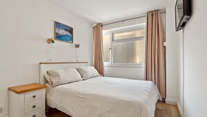 A bedroom with a bed, a nightstand, a TV mounted on the wall, a window with curtains, and a painting on the wall. - Life's a Beach - Direct Beach Access, Sea Views (Saundersfoot)