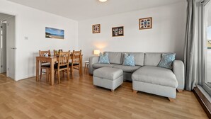 A living room with a grey sofa, wooden dining table, and chairs. There are framed pictures on the wall and a window with curtains. - Life's a Beach - Direct Beach Access, Sea Views (Saundersfoot)