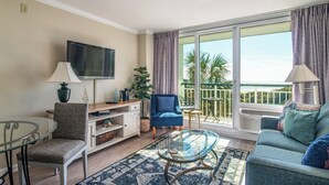 A living room with a TV, a blue chair, a glass coffee table, and a view of palm trees through large windows.