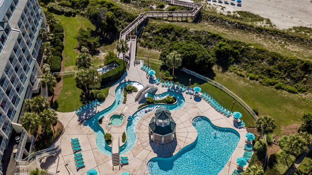 An aerial view of a resort with multiple swimming pools, a central water feature, and a surrounding deck area with lounge chairs and umbrellas.