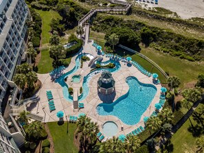 An aerial view of a resort with multiple swimming pools, a central water feature, and a surrounding deck area with lounge chairs and umbrellas.