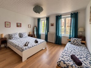 Quarto com uma cama grande, uma cama menor, um espelho, um radiador e uma janela com cortinas.