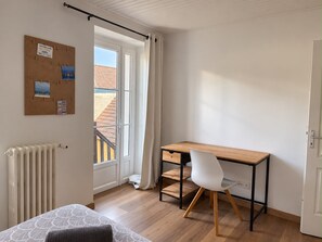 Une pièce avec un bureau en bois, une chaise blanche, un radiateur, un tableau d’affichage en liège où sont accrochées des photos, et une fenêtre avec des rideaux.