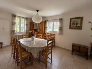 Uma sala de jantar com mesa redonda, cadeiras de madeira, um vaso com flores, luminária pendente e um quadro na parede.