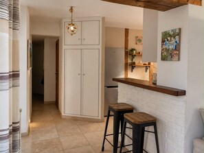 Cozinha com parede de tijolos brancos, bancada de madeira, dois banquinhos, luminária pendente e geladeira.