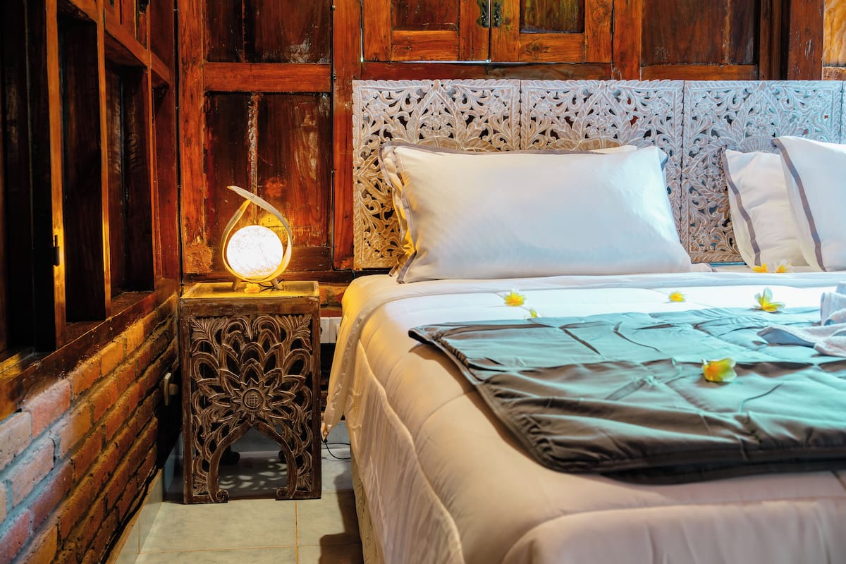 Una cama bien hecha con sábanas blancas y una manta verde, una mesita de noche de madera con una lámpara decorativa, y una pared con intrincados diseños de azulejos.