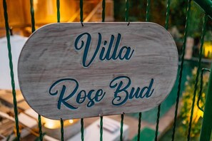 Ein hölzernes Schild mit der Aufschrift 'Villa Rose Bud', an einem Metallzaun befestigt.
