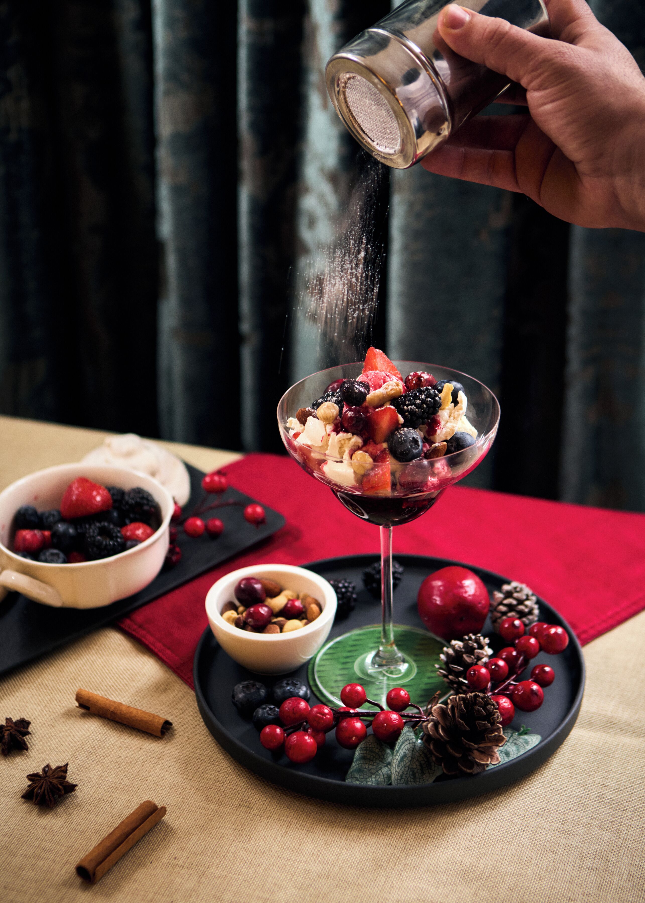 A dessert in a glass with a variety of fruits and nuts, a bowl of berries, and a plate with pinecones and berries.