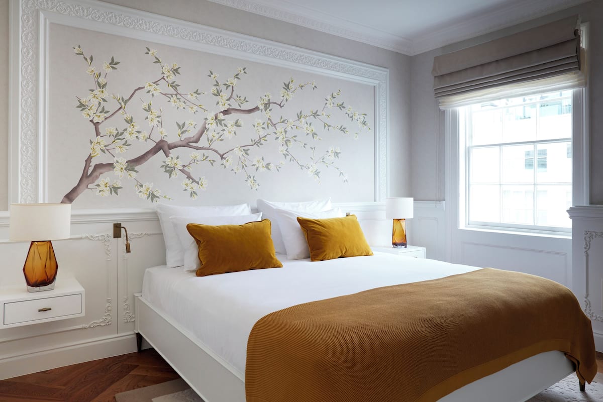 Un lit bien fait, avec une literie blanche et moutarde, un tableau représentant un arbre accroché au mur, et une fenêtre avec des rideaux.
