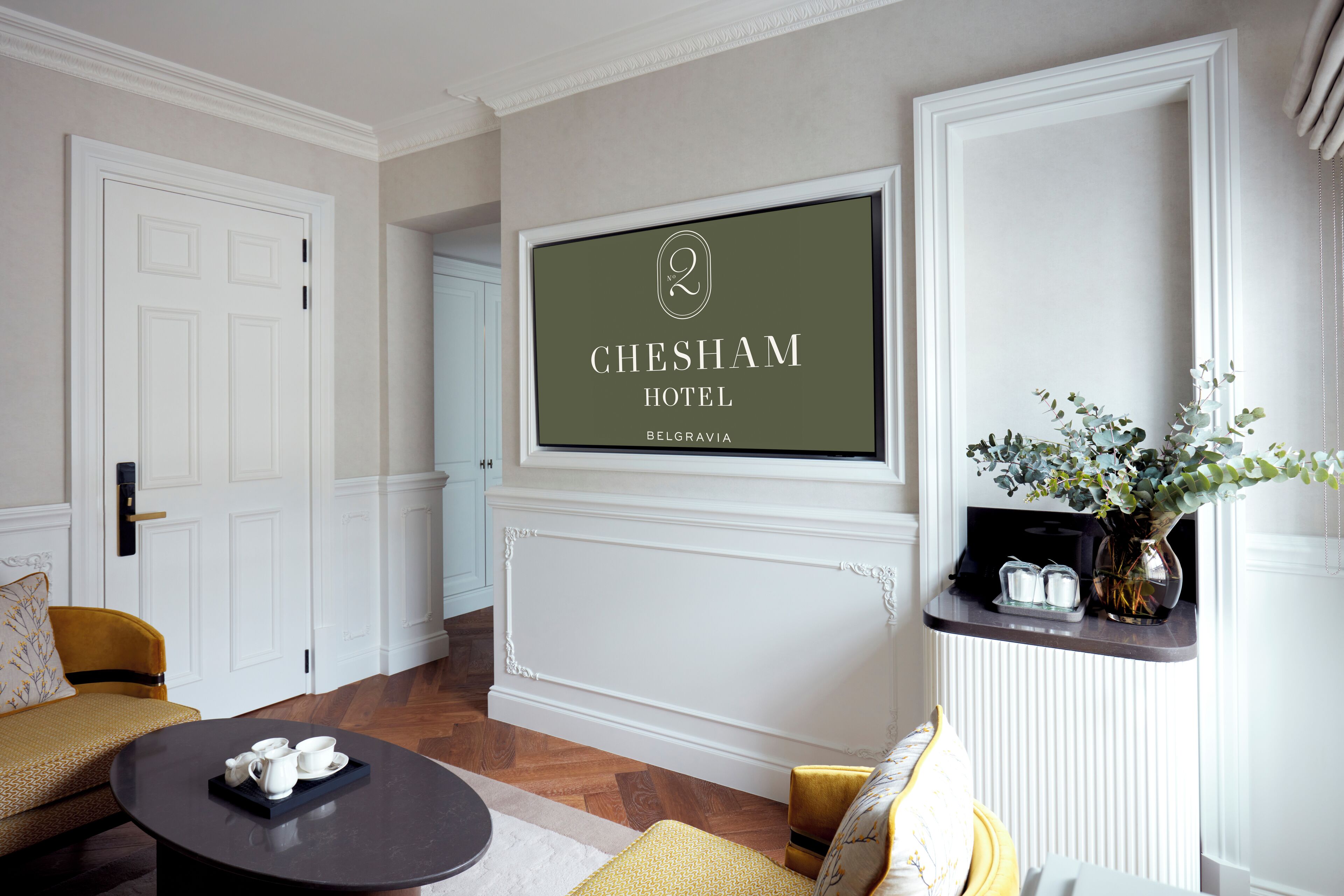 A hotel lobby with a reception desk, a sofa, a coffee table with tea set, and a wall-mounted sign for Chesham Hotel.