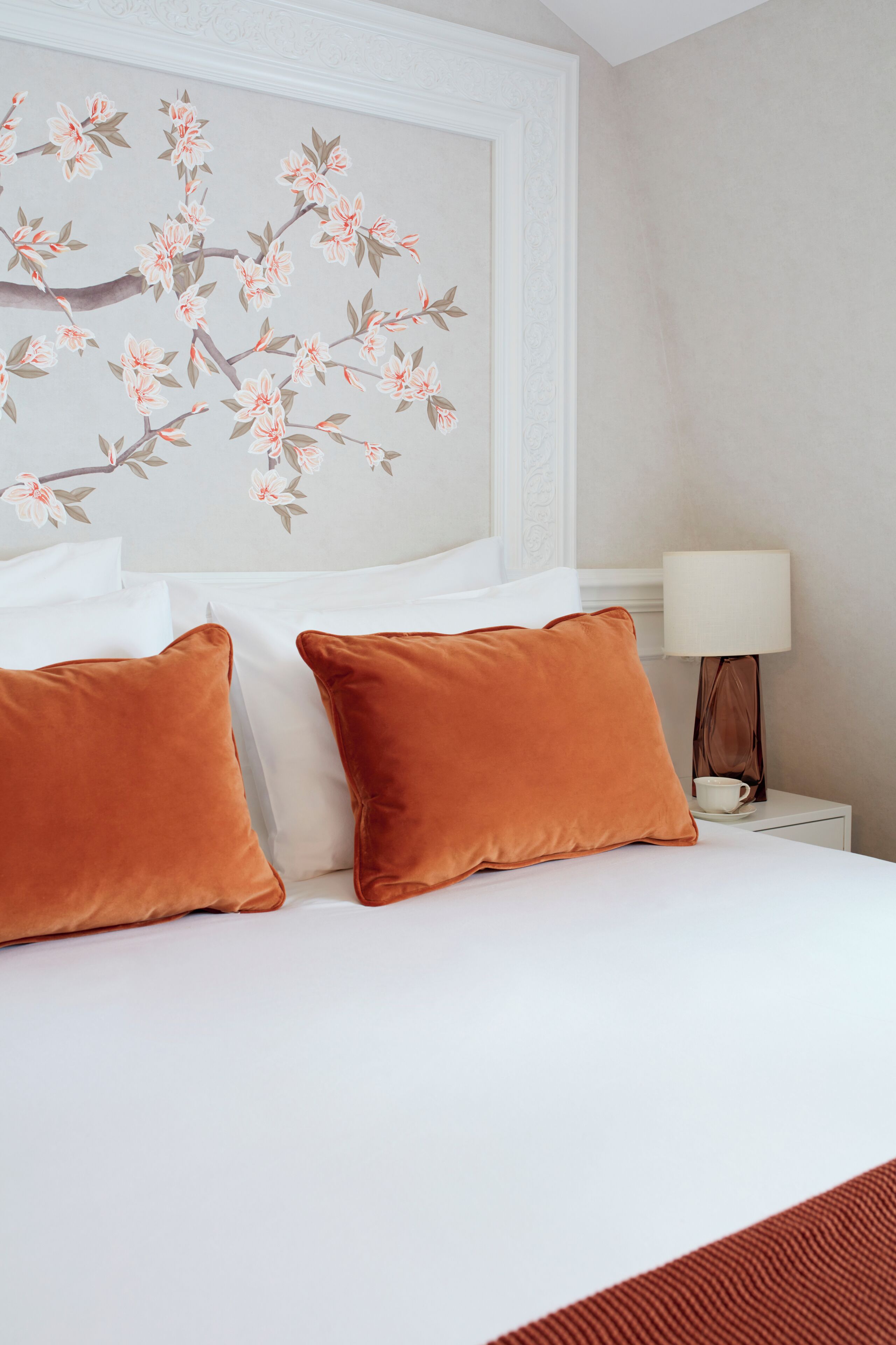 Un lit bien fait, agrémenté d’oreillers blancs et orange, un mur décoré d’un motif floral et une table de chevet avec une lampe et une tasse.