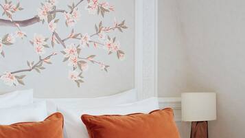Un lit bien fait, agrémenté d’oreillers blancs et orange, un mur décoré d’un motif floral et une table de chevet avec une lampe et une tasse.