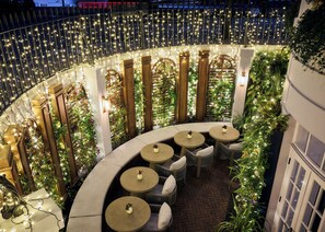 A circular outdoor seating area with round tables and chairs, surrounded by a decorative railing with string lights. - 2 Chesham Hotel (London)