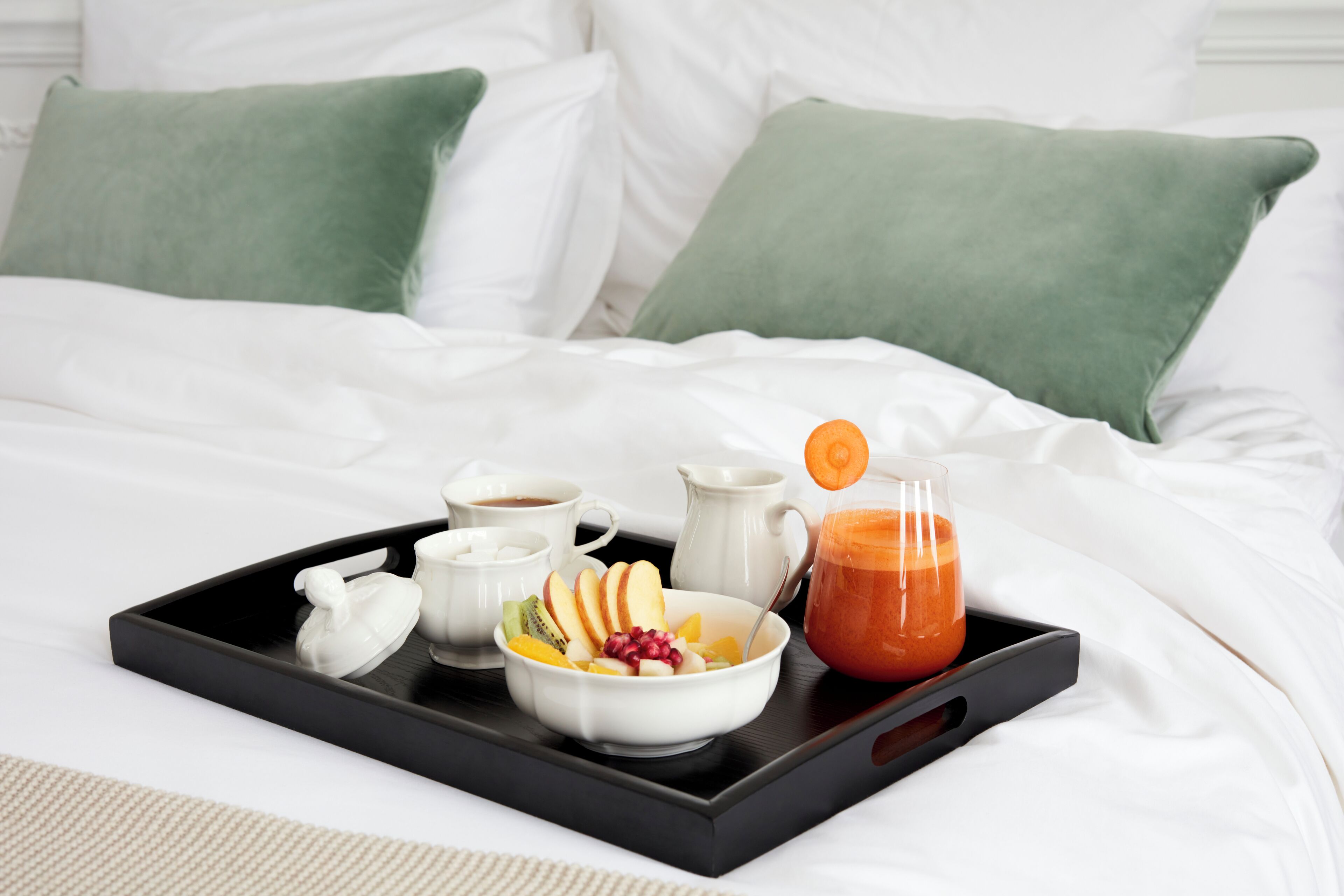 A breakfast tray with fruits, a glass of orange juice, and a coffee cup on a bed with white bedding and green pillows.