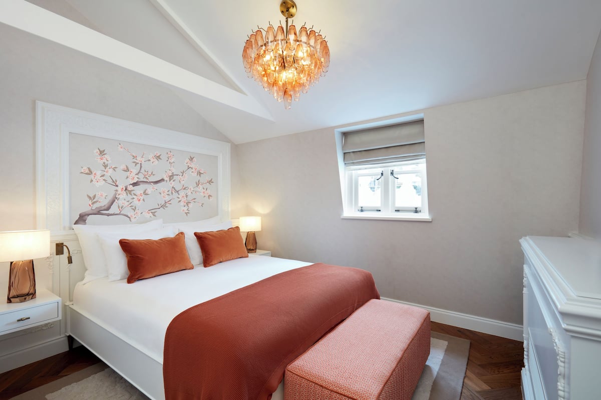 Une chambre à coucher avec un grand lit, un lustre, une fenêtre avec des stores et une table de chevet avec une lampe.