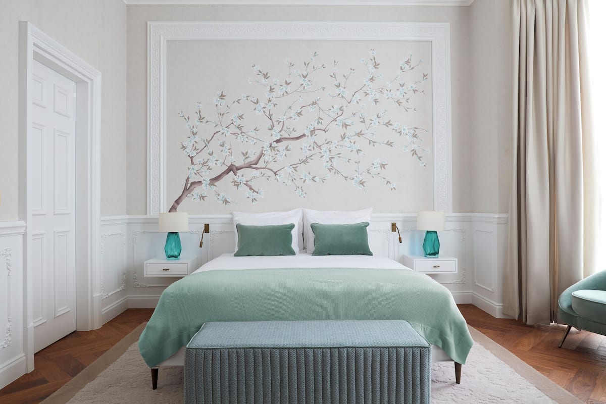 Une chambre avec un grand lit, deux tables de chevet avec des lampes, un banc recouvert d’un tissu vert et une œuvre d’art représentant une branche d  ’arbre, accrochée au mur.