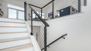Una escalera moderna con escalones de madera y barandillas de metal negro, que conduce a una zona del segundo piso con vista al cielo.