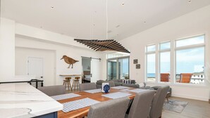Un comedor espacioso con una mesa y sillas de madera, una cocina al fondo y ventanas amplias con vista al mar.