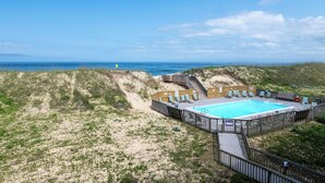 Et poolområde ved stranden med træterrasse og liggestole, omgivet af klitter og hav.