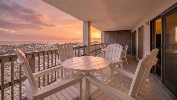 En balkon med et rundt bord og fire Adirondack-stole, med udsigt over en strand ved solnedgang.