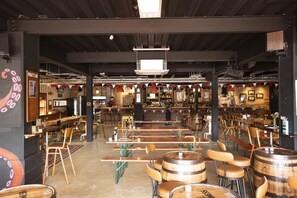 L’intérieur d’un restaurant est aménagé avec des tables et des chaises en bois, un coin bar et un plafond haut avec des poutres apparentes.