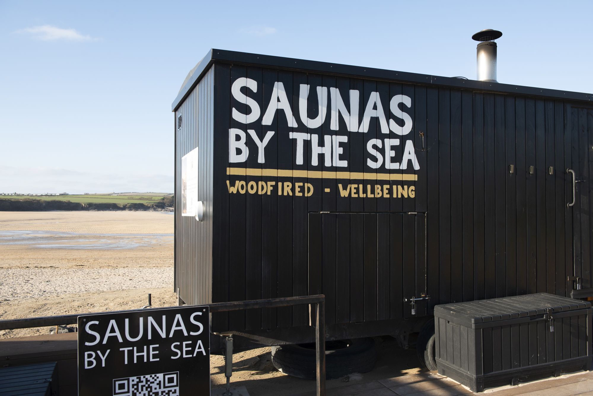 A sauna structure labeled "SAUNAS BY THE SEA" with a sign indicating "WOODFIRED - WELLBEING".