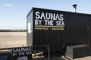 Une structure de sauna portant l’inscription « SAUNAS BY THE SEA » et affichant un panneau indiquant « CHAUFFÉ AU BOIS – BIEN-ÊTRE ».